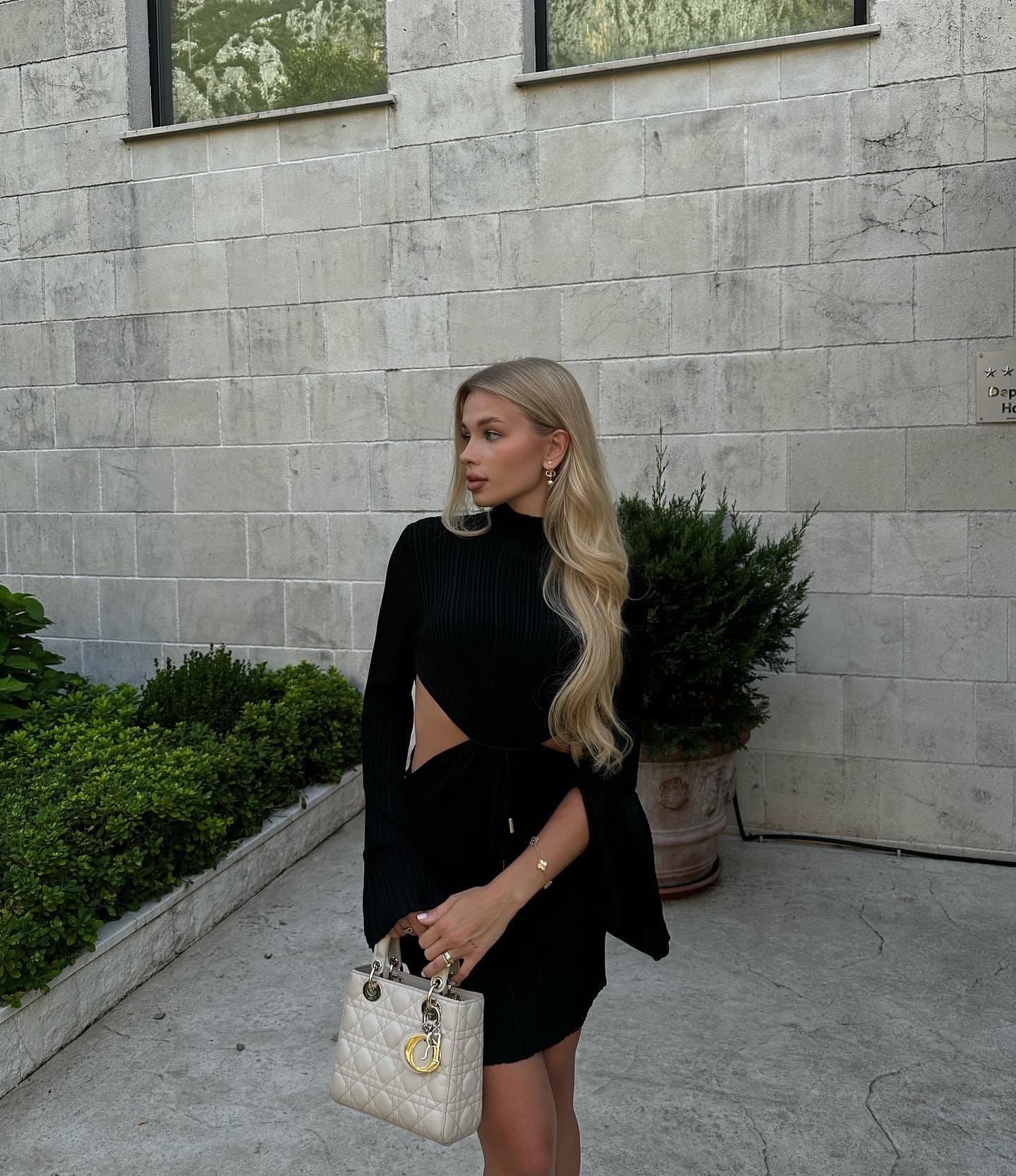A blonde woman stands in profile against a grey stone wall, wearing a black ribbed cutout top and skirt with a white handbag.