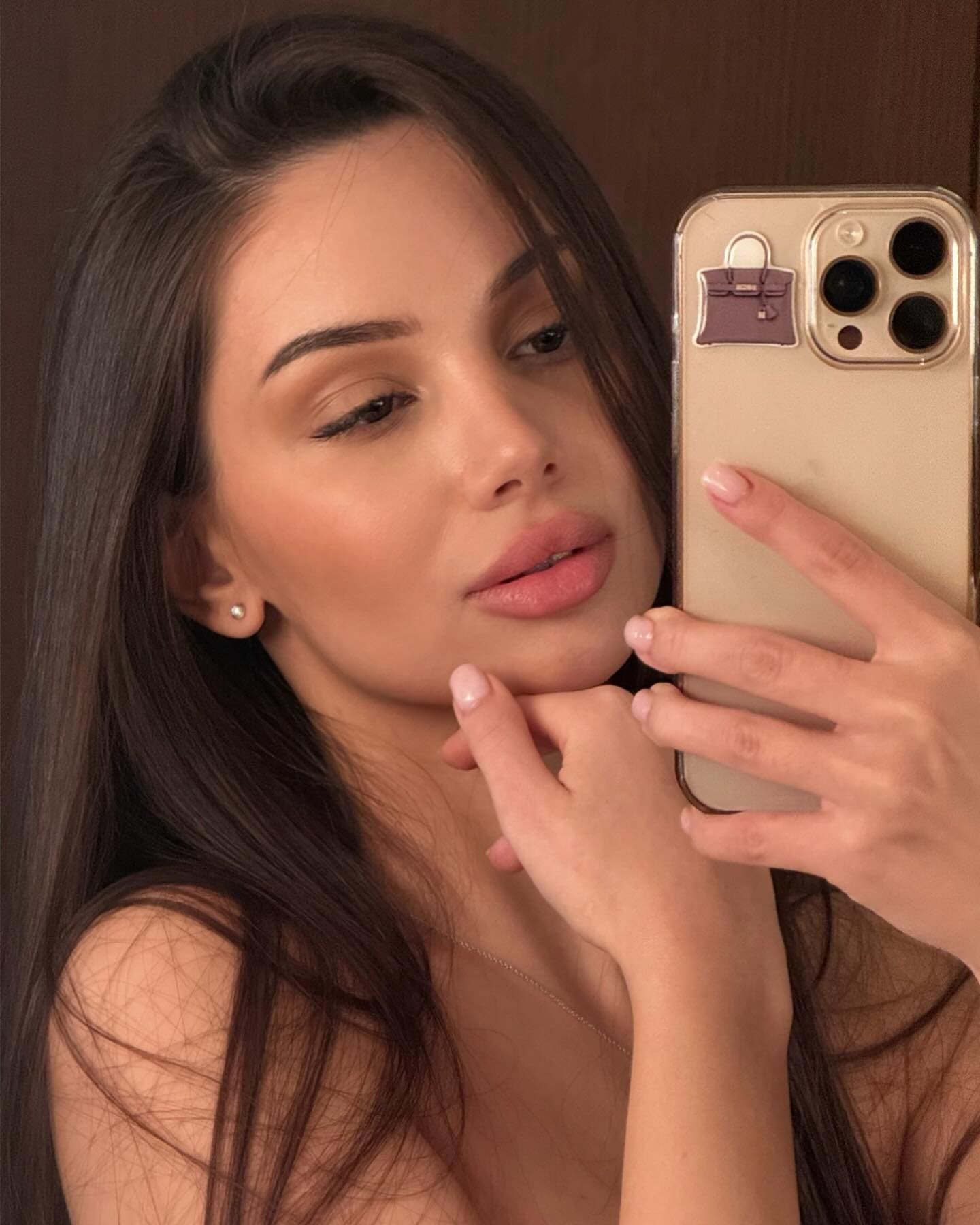 Mirror selfie in a sheer top with hand under chin, holding a gold phone against a brown background.
