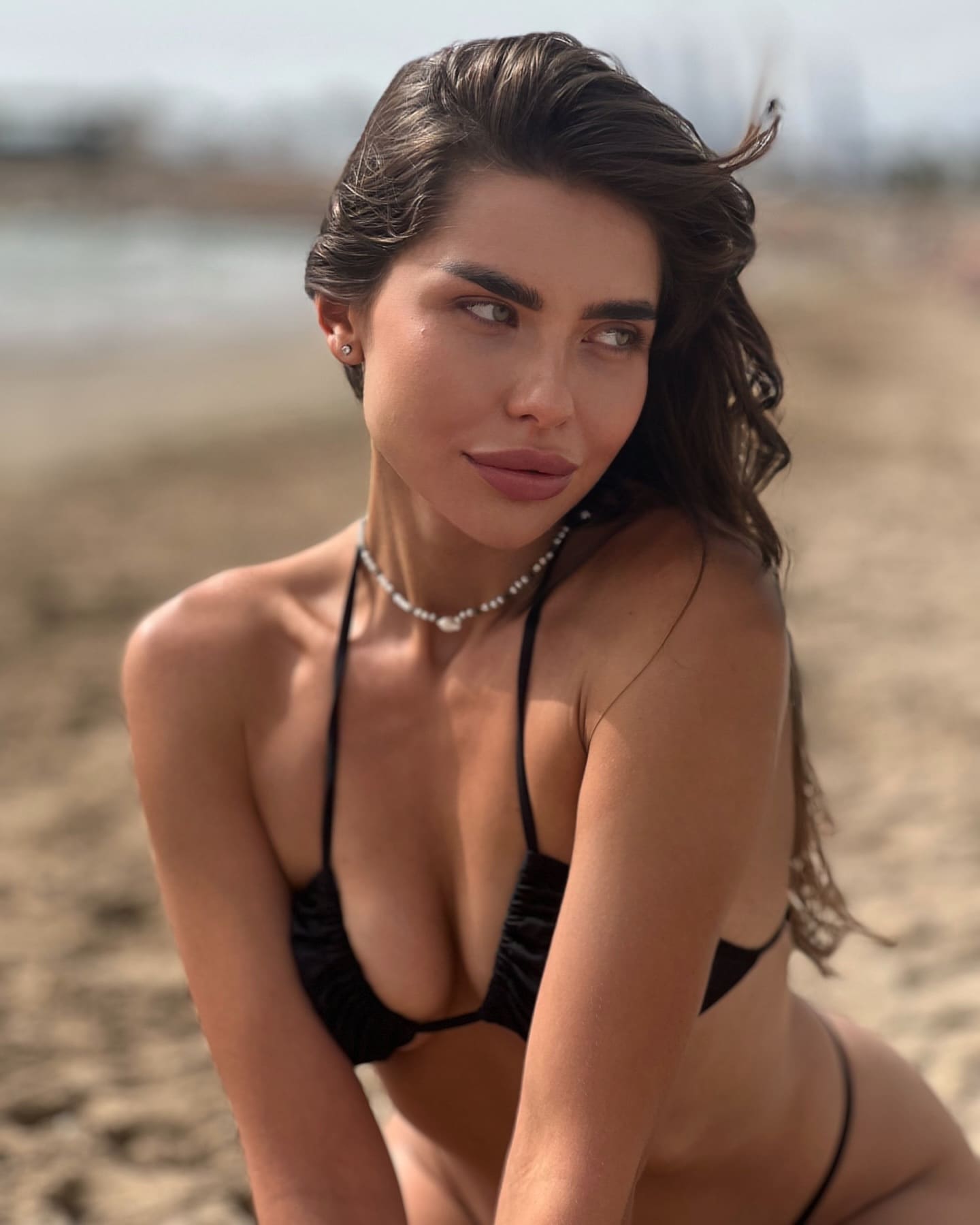 A woman with long dark hair sits on a sandy beach in a black string bikini and beaded necklace, looking to the side.
