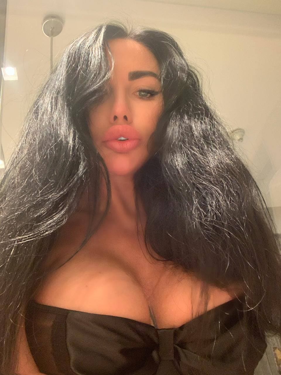 A close-up mirror selfie shows a woman in a black strapless top with a deep neckline, taken in a bright room with a shower visible.