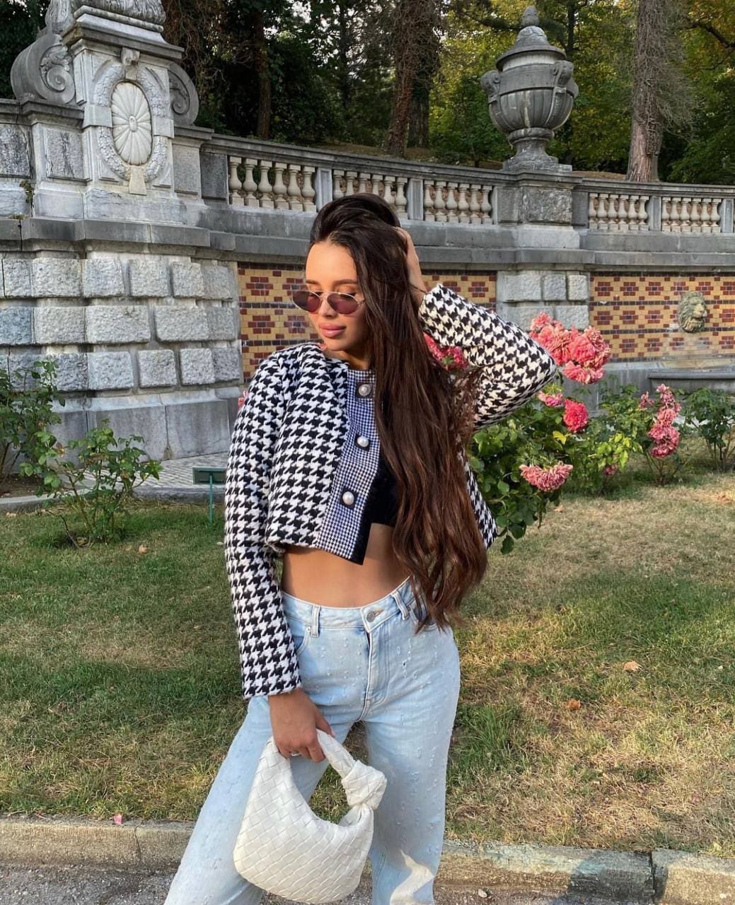 A woman stands in a garden wearing a black-and-white houndstooth cropped jacket and light-wash jeans, holding a white handbag near a stone balustrade.