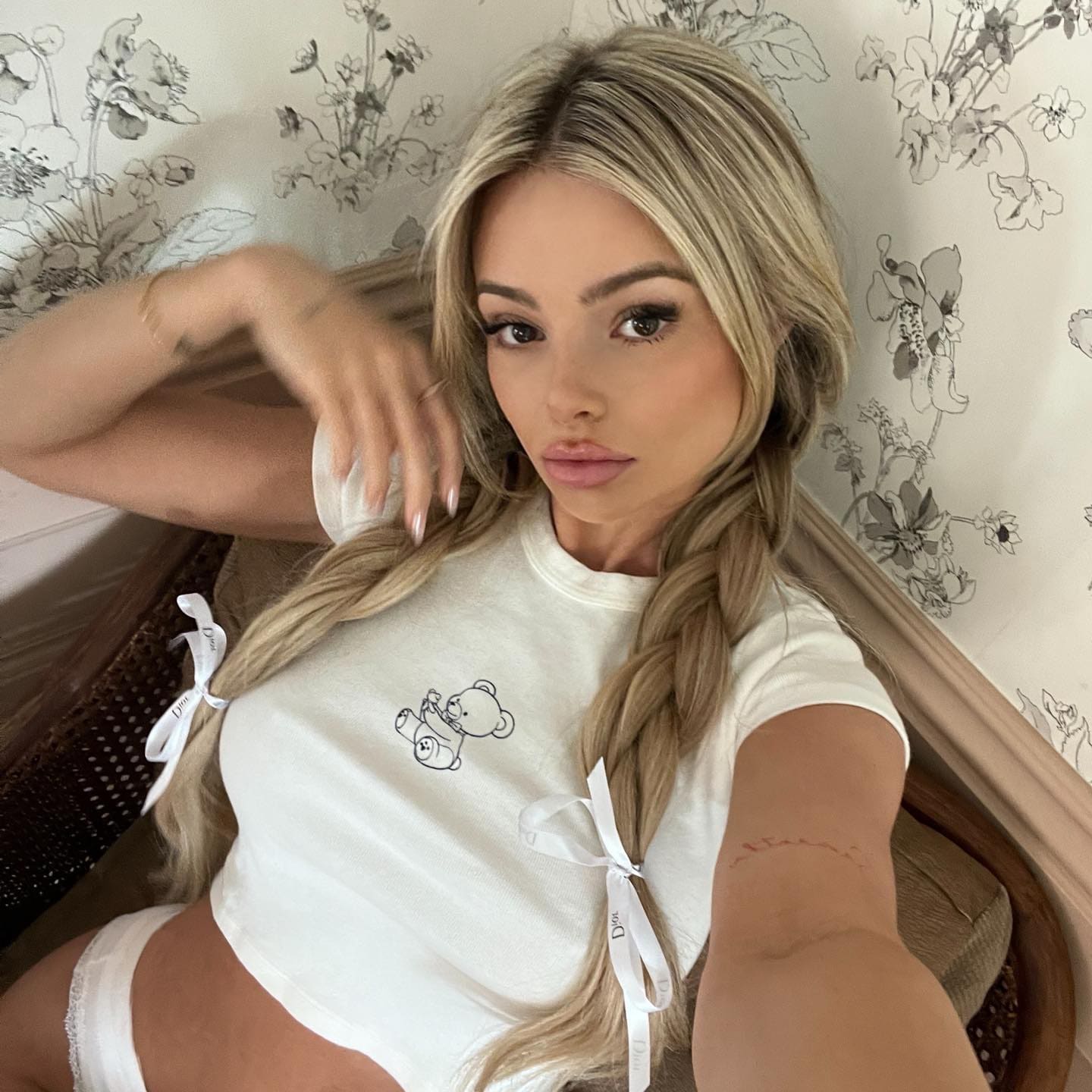 A blonde woman with braided hair and white ribbons sits against floral wallpaper, wearing a white t-shirt with a bear graphic.